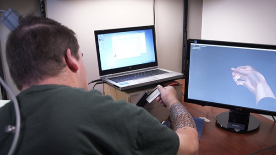 Descrição da imagem: Homem aparece de costas, em frente a dois computadores. Ele retira um cartão da carteira com a mão direita.