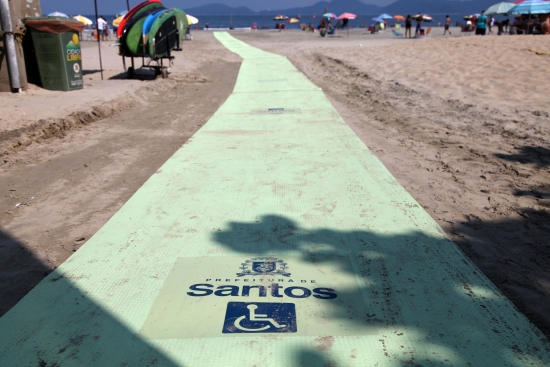 Descrição da Imagem: esteira acessível na cor branca está colocada sobre a faixa de areia e vai até o mar. No equipamento estão impressos o brasão e o nome da cidade de Santos, e também o ícone da cadeira de rodas que simboliza a acessibilidade.