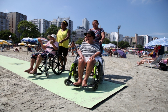 Descrição da Imagem: Duas cuidadoras estão paradas atrás da cadeiras onde suas clientes estão sentadas. Todas estão sobre a esteira acessível na praia de Santos. Ao fundo, pessoas sentadas em cadeiras de praia e prédios da cidade.