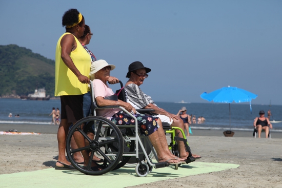 Descrição da imagem: No primeiro plano, duas senhoras estão sentadas em suas cadeiras de rodas, que são empurradas por cuidadoras na praia de Santos, sobre as esteiras acessíveis. Ao fundo, o mar e o céu aparecem em uma cor azul.