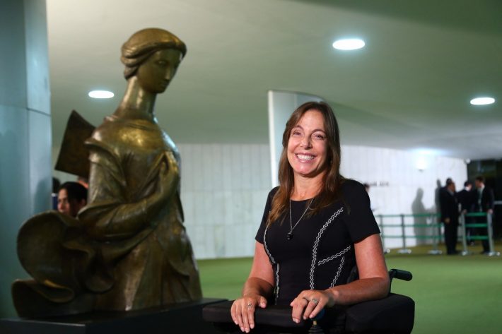 Descrição da imagem #pracegover: Mara Gabrilli está no salão verde da Câmara dos Deputados, em Brasília. Usa vestido preto e sorri. Ao seu lado direito está a escultura de um anjo, com a mão no peito, que parece olhar para ela. Foto: Divulgação
