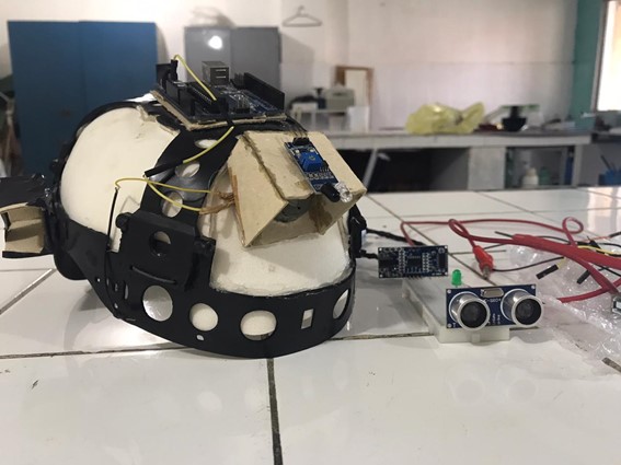 Foto de um capacete branco de construção civil adaptado com sensor de movimentos. Em volta estão peças de papel paraná e EVA,, bateria, cabo de alimentação e outros itens como uma placa eletrônica. Crédito: Divulgação.