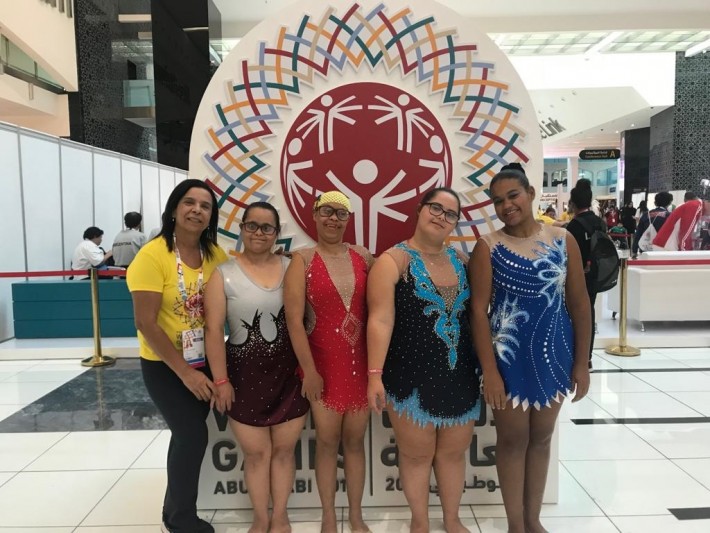 Foto de cinco mulheres que estão em pé na frente da logomarca da 'Special Olympics'. Registro feito no ano passado em Abu Dhabi, nos Emirados Árabes Unidos. Crédito: Divulgação.