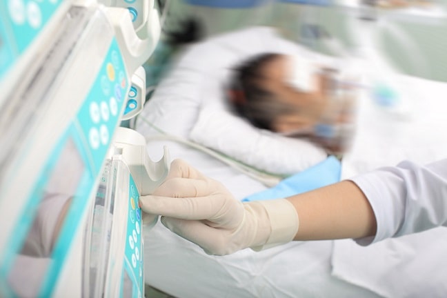 Foto de uma pessoa intubada e deitada em leito hospitalar. À frente, um profissional de saúde manuseia equipamentos. Crédito: Reprodução.