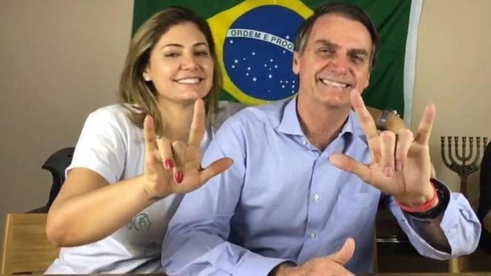 Foto da primeira-dama Michelle Bolsonaro e do presidente Jair Bolsonaro juntos, olhando para a câmera e sorrindo, fazendo 'i love you' (eu amo você) com as mãos na língua de sinais. Crédito: Reprodução.