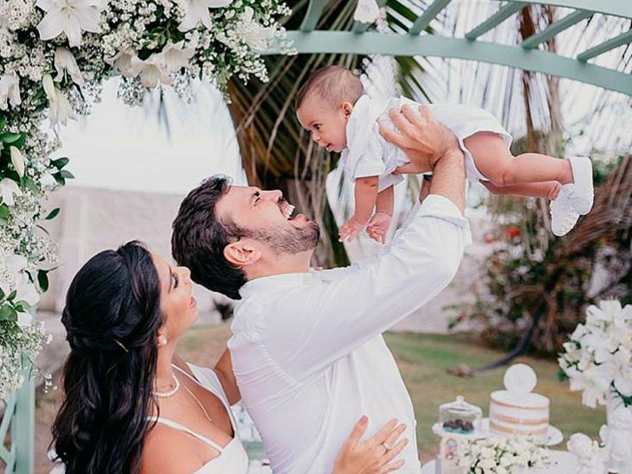 Foto de Fabíola e Igor com o bebê Miguel. Os três vestem roupas brancas. Igor eleva Miguel com os braços e está sorrindo. Ao redor, flores brancas. Crédito: Divulgação.