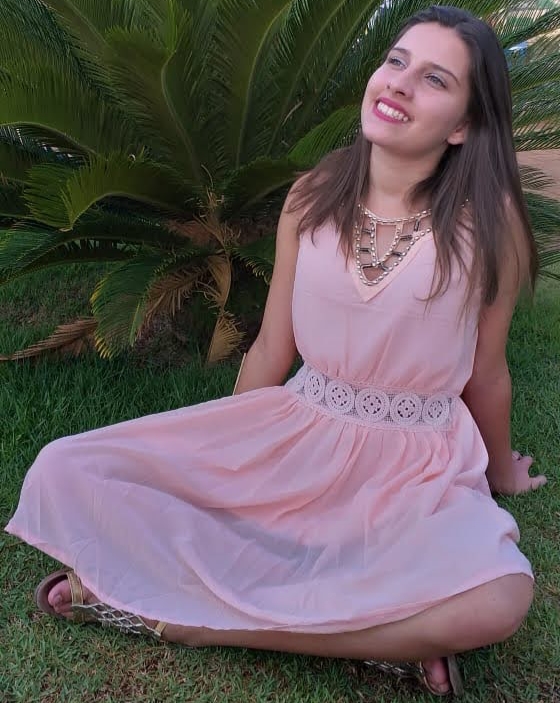 Foto de Jenifer Lemos antes das sequelas da doença de Lafora. Ela está sentada em um gramado, com as pernas cruzadas, olhando para o alto e sorrindo. Usa um vestido rosa. Crédito: Arquivo pessoal / Laci Lemos.