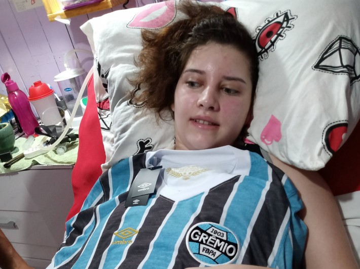 Foto de Jenifer Lemos deitada na cama. A adolescente tem a pele clara, cabelos e olhos castanhos. Está coberta por uma camisa do time de futebol Grêmio. Crédito: Arquivo pessoal / Laci Lemos.