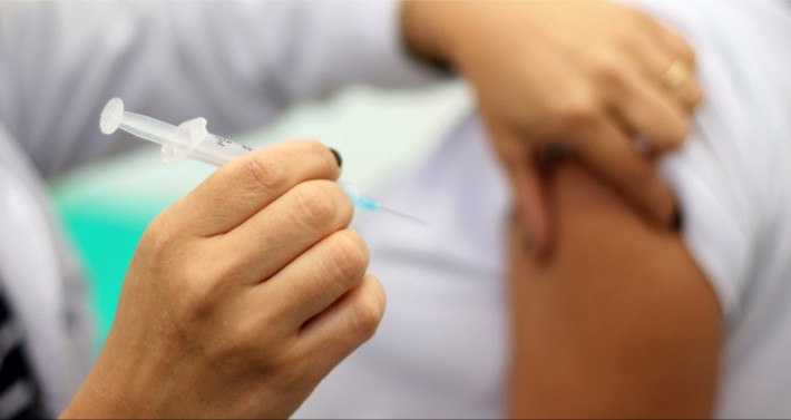 Foto das mãos de uma profissional de saúde segurando uma seringa com agulha e apertando o braço de uma pessoa na altura do ombro para injeção de medicamento. Crédito: Reprodução.