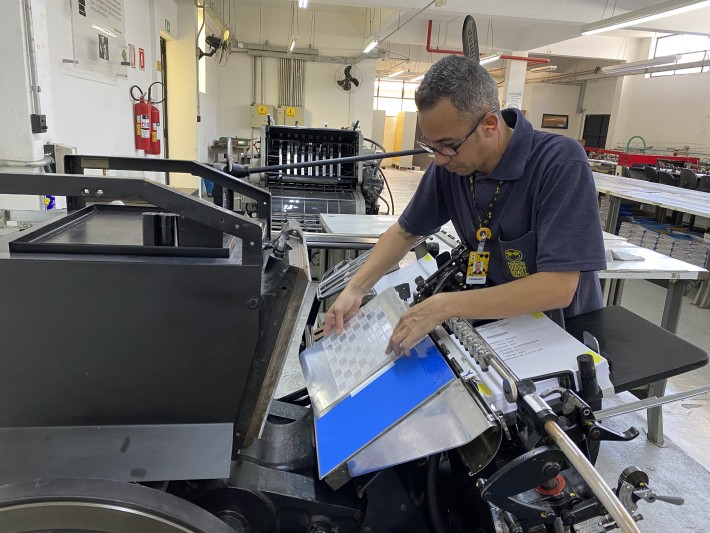 Foto da gráfica em Braille da Fundação Dorina Nowill para Cegos mostra um funcionário operando uma impressora manual e inserindo no equipamento uma placa de alumínio. Crédito: Divulgação.