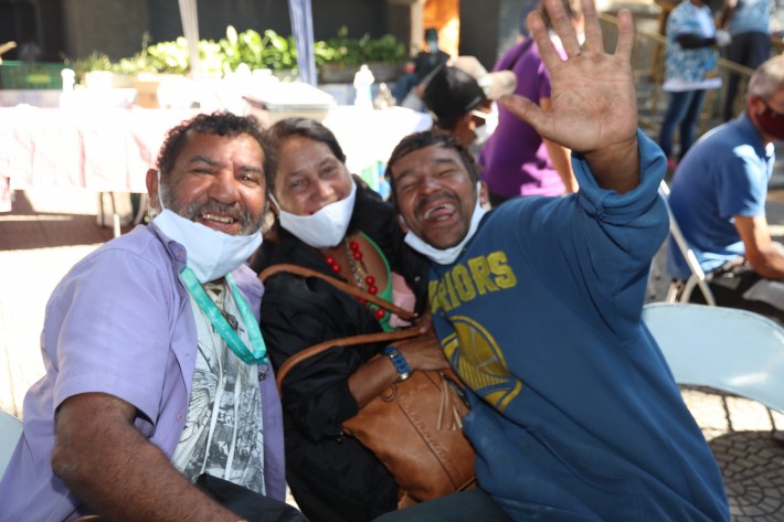 Foto de uma mulher e dois homens, que estão abraçados, sorrindo e acenando para a câmera. Crédito: Monica Zanon / Divulgação.