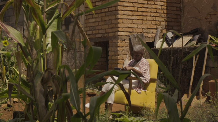 Foto aberta que mostra a cantora Luiza da Iola sentada em uma poltrona amarela, folheando um álbum de fotografias. Ao redor, vegetações e uma construção de tijolos aparentes.