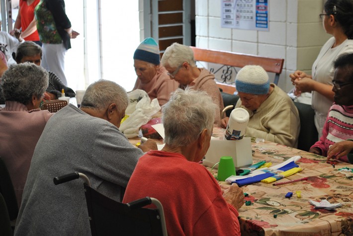 Foto de um grupo de idosos, homens e mulheres, em volta de uma mesa, fazendo artesanato. Alguns usam casacos e gorros, outras estão em cadeiras de rodas. Em volta, pessoas mais novas estão em pé.