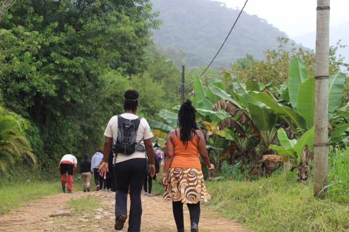 Foto de várias pessoas negras caminhando por uma estrada de terra, no meio de uma ampla vegetação.