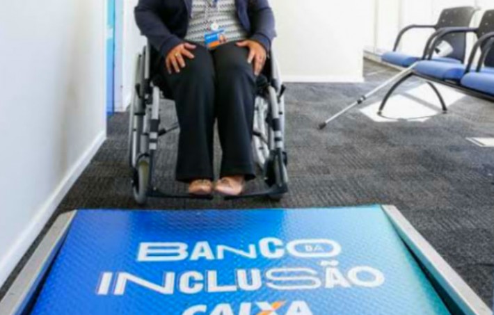 Foto de mulher em cadeira de rodas, dentro de uma unidade da Caixa Econômica Federal. Na frente da mulher, uma rampa com a frase 'banco inclusão caixa'.