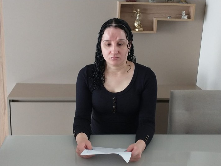 Foto de Helena Rosa Gois, mulher branca, de 29 anos, com cabelos escuros, ondulados e compridos. Ela tem o semblante sério e está sentada à mesa segurando um documento. Ao redor, alguns móveis e objetos de decoração.