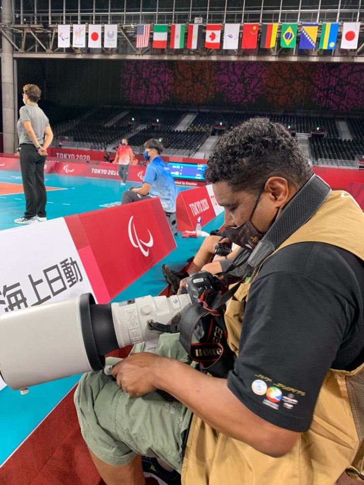 Foto de João Maia na arquibancada de um ginásio onde é realizada uma competição de Judô. Ela segura uma câmera com uma grande lente, enquanto configura o equipamento. Ao fundo, bandeiras de diversos países penduradas.