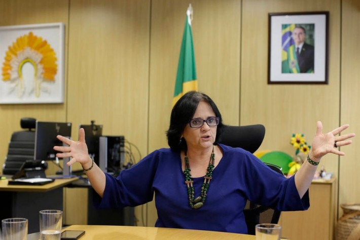 Foto da ministra Damares Alves em seu gabinete. Ela está com os braços abertos e falando.