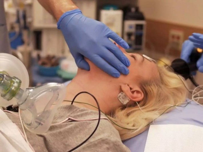 Foto de uma mulher de cabelos loiros deitada em um leito hospitalar, descordada, sendo submetida a uma eletroconvulsoterapia.