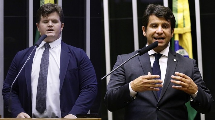 Foto dupla. Na esquerda, o deputado Felipe Rigoni e na direita o deputado Dr. Luizinho. Ambos são brancos, veste, ternos escuros e discursam no plenário da Câmara.