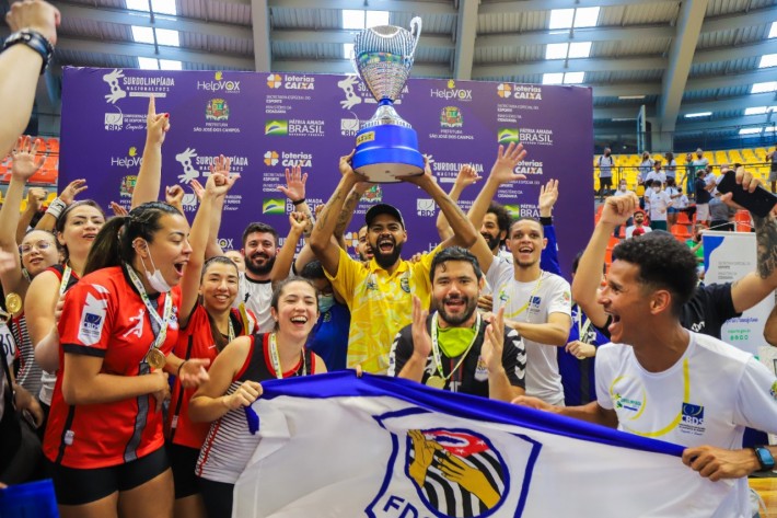 Equipe da Federação de São Paulo comemora a vitória na Surdolimpíada Nacional. Grupo exibe o troféu da competição e balança a bandeira do time.