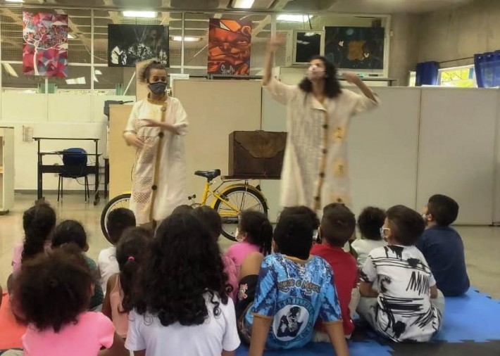 Encenação do espetáculo 'Histórias Gregas em Sinais e Imagens' em uma sala. Duas atrizes se apresentam para um grupo de crianças. Atrás da artistas está a bicicleta com a caixa-cenário.