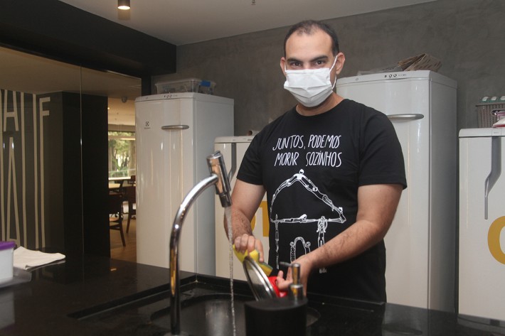Foto de Nicolas Munhoz, de 30 anos, homem branco, que está em pé na frente da pia de uma cozinha, lavando louça. Ele veste uma camisa preta com a frase 'juntos, podemos morar sozinhos'.