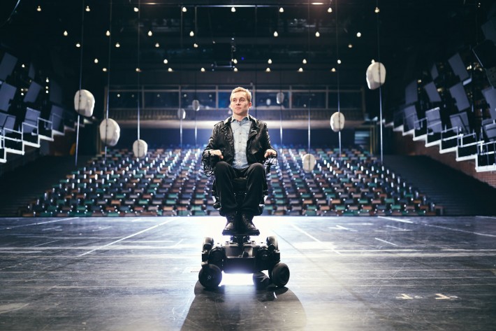 Foto de Samuel Koch, ator alemão de 34 anos, branco, de cabelos claros e curtos. Ele é tetraplégico, está sentado em uma cadeira de rodas motorizada, sobre um palco, de costas para a plateia vazia. As luzes do teatro estão acesas.