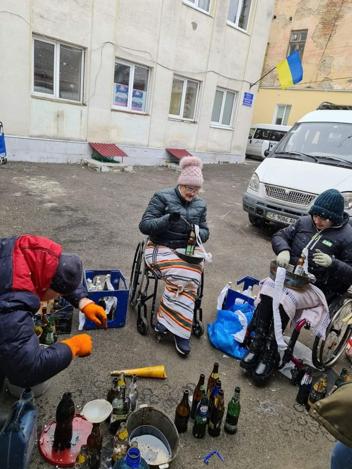 Foto inteira, feita na posição vertical, mostra um prédio com a bandeira da Ucrânia pendurada, e três pessoas na área externa, com piso de terra. Um homem e duas mulheres estão montando coquetéis molotov. As duas mulheres, uma idosa e uma menina, estão em cadeiras de rodas.