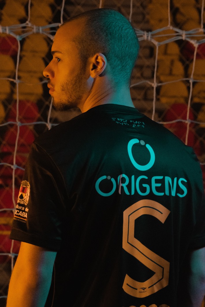 Foto de homem negro, de costas para a câmera, vestindo uniforme escuro do time de futsal Magnus. No alto da camisa, uma impressão em braile próxima ao colarinho traz a mensagem 'Magnus Futsal desde 2014'.