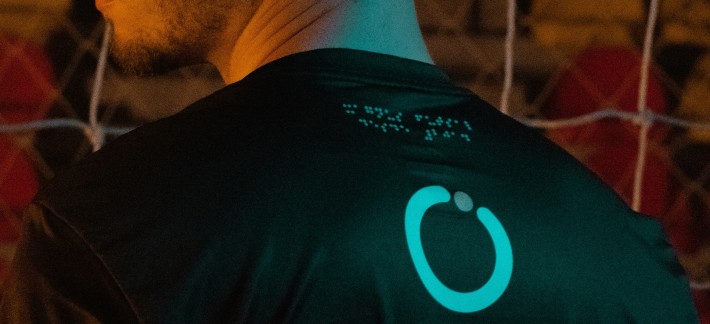 Foto de homem negro, de costas para a câmera, vestindo uniforme escuro do time de futsal Magnus. No alto da camisa, uma impressão em braile próxima ao colarinho traz a mensagem 'Magnus Futsal desde 2014'.