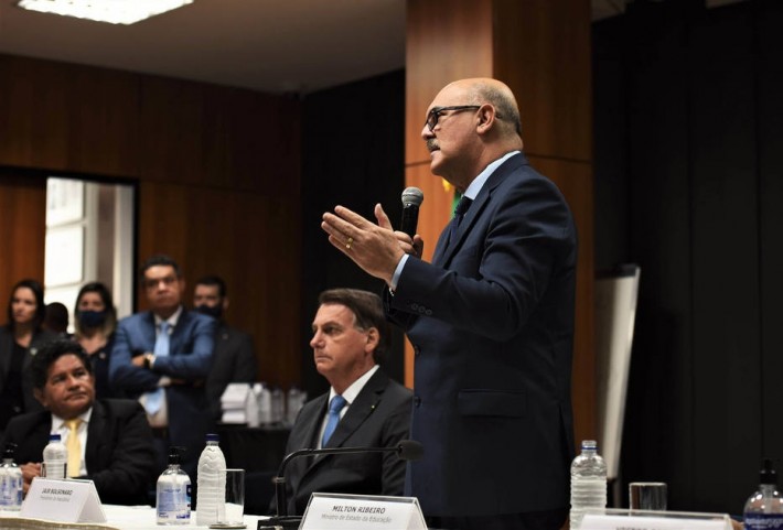 Foto do ministro da Educação, MIlton Ribeiro, em pé, falando ao microfone, ao lado do presidente da República, Jair Bolsonaro, que está sentado.