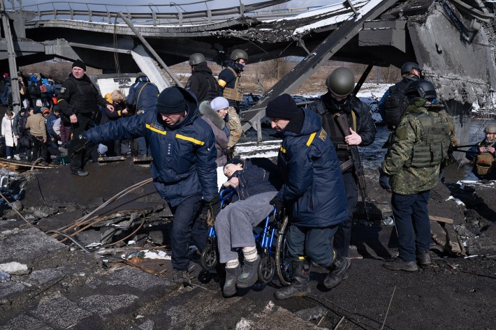 Foto de uma mulher em cadeira de rodas sendo carregada por dois homens em meio a uma área destruída na Ucrânia.