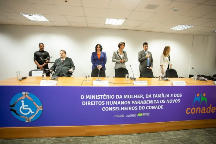 Foto da cerimônia de posse do Conade, 2022/2025. Damares Alves, Michelle Bolsonaro e Cláudio Panoeiro estão em pé ao lado de outras pessoas com e sem deficiência.