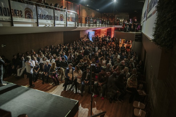 Foto de um grande grupo de pessoas sentadas em frente a um palco onde um homem faz uma apresentação.