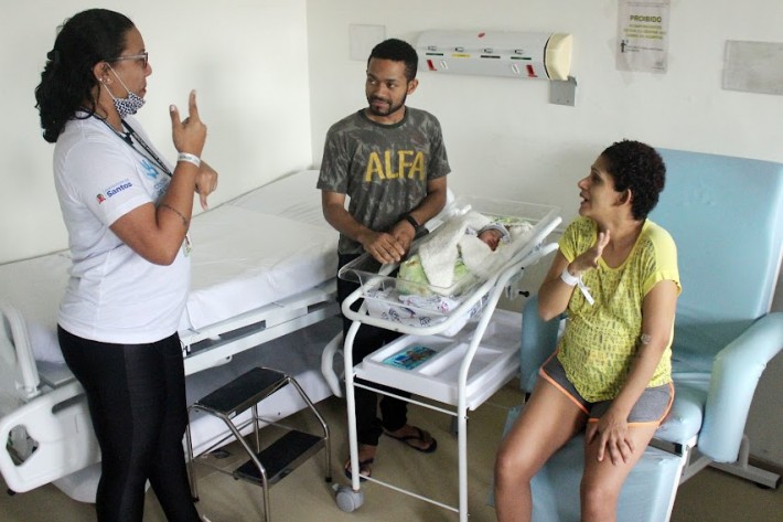 Foto de três pessoas, duas mulheres e um homem, conversando em Libras no quarto de um hospital. Um bebê repousa num berço.